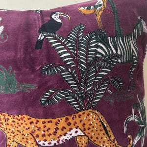 Op de afbeelding: Zacht, paars kussen met een jungle-thema ontwerp. Het kussen heeft een cheetah, zebra, toekan en weelderig groen gebladerte. De cheetah heeft een oranje en bruin gevlekt patroon. De zebra heeft zwart-witte strepen.