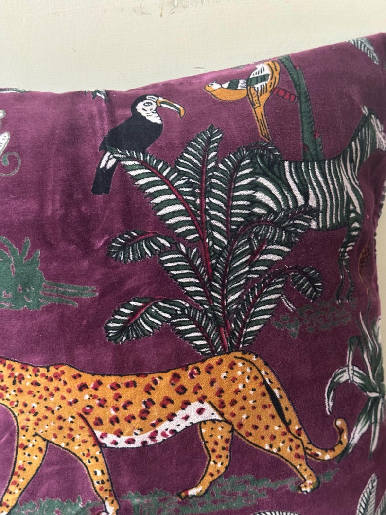 Op de afbeelding: Zacht, paars kussen met een jungle-thema ontwerp. Het kussen heeft een cheetah, zebra, toekan en weelderig groen gebladerte. De cheetah heeft een oranje en bruin gevlekt patroon. De zebra heeft zwart-witte strepen.