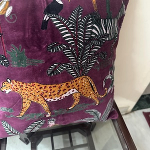 Op de afbeelding: Vierkant kussen in dieppaars. Het heeft een jungle-thema print met een luipaard, toekans en een zebra. Het ontwerp omvat weelderig groen gebladerte op een rijke, fluweelzachte achtergrond.