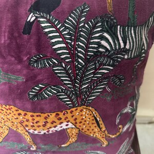 Op de afbeelding: Bordeaux fluwelen kussen met een junglethema. Voorzien van een lopende luipaard met oranje vlekken, een zebra, toekan en andere vogels. Het kussen heeft een palmboomontwerp met groene en witte bladeren.