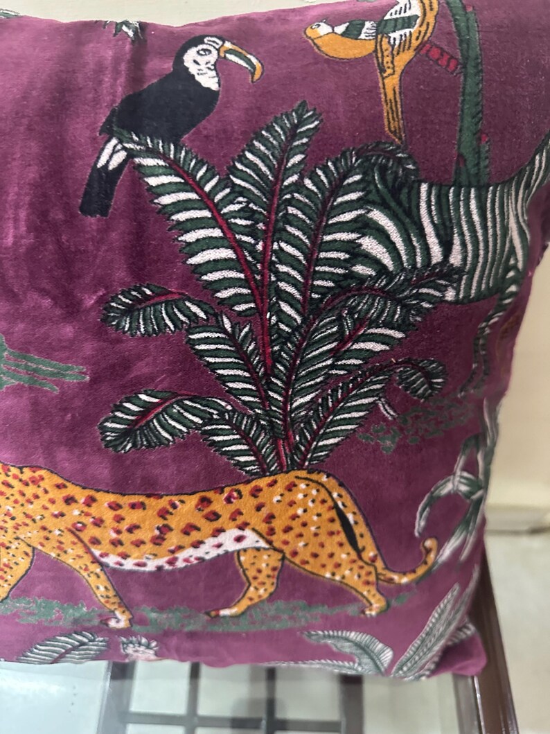 Op de afbeelding: Bordeaux fluwelen kussen met een junglethema. Voorzien van een lopende luipaard met oranje vlekken, een zebra, toekan en andere vogels. Het kussen heeft een palmboomontwerp met groene en witte bladeren.
