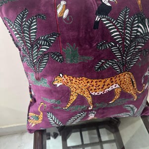 Op de afbeelding: Zacht, pruimkleurig decoratief kussen met een junglesc&egrave;ne. Het ontwerp omvat een luipaard, apen, toekans en palmbomen in tinten oranje, wit en groen. Het kussen ligt op een donkere houten tafel.