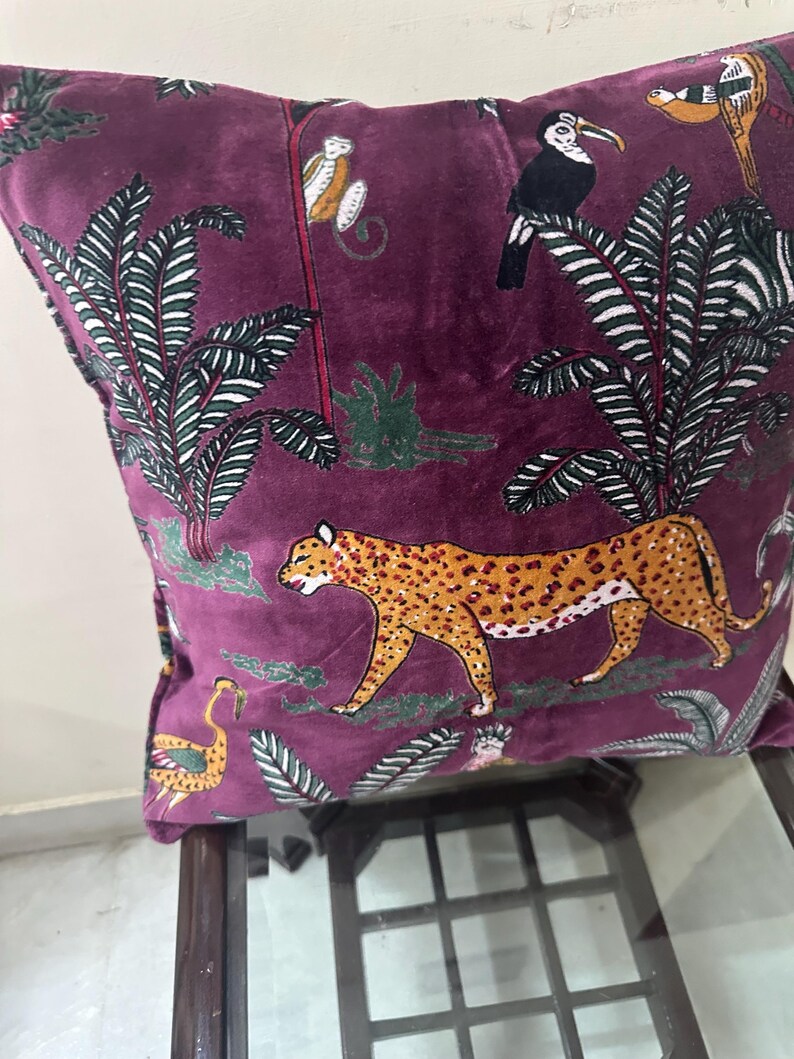 Op de afbeelding: Zacht, pruimkleurig decoratief kussen met een junglesc&egrave;ne. Het ontwerp omvat een luipaard, apen, toekans en palmbomen in tinten oranje, wit en groen. Het kussen ligt op een donkere houten tafel.