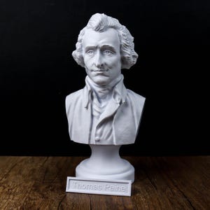May include: A white bust of Thomas Paine, a historical figure, with detailed facial features and curly hair. The bust is on a pedestal, with a nameplate that reads "Thomas Paine". The sculpture is set against a dark background.