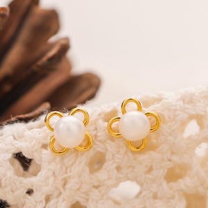 May include: A pair of gold-colored flower-shaped stud earrings with a white pearl center. The earrings are displayed on a white, textured surface. The brand name "YAMMY JEWELRY" is visible in the lower part of the image.
