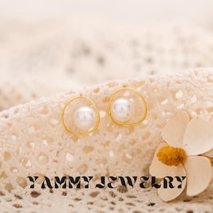 Puede incluir: Un par de pendientes dorados, cada uno con una perla blanca redonda y grande, engastada en un delicado marco circular abierto. Los pendientes descansan sobre una superficie texturizada de color crema. Se ve el nombre de la marca "YAMMY JEWELRY".