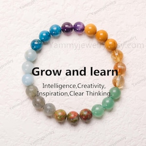 Puede incluir: Un brazalete de cuentas de colores con piedras en tonos azules, verdes, amarillos, naranjas, morados y grises. El brazalete tiene el texto "Grow and learn" y "Intelligence, Creativity, Inspiration, Clear Thinking" impreso sobre un fondo blanco.