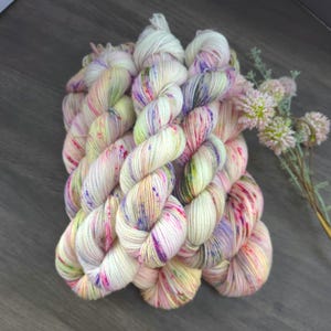 May include: A bundle of hand-dyed yarn in cream, pink, purple, and green hues. The yarn is twisted into skeins, with a speckled appearance. A sprig of pink flowers is in the background.