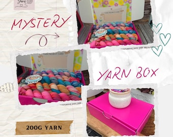 Puede incluir: Una caja de hilo rosa con el texto "YARN BOX" y "MYSTERY". Dentro de la caja hay madejas de hilo coloridas. La imagen también muestra una etiqueta de hilo de 200 g y un tarro pequeño.