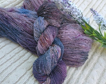 Puede incluir: Una madeja de hilo teñido a mano con una paleta de colores variados en tonos morado intenso, rosa empolvado y azul pizarra. El hilo tiene una textura ligeramente irregular y se muestra junto a una rama de pequeñas flores blancas y moradas.