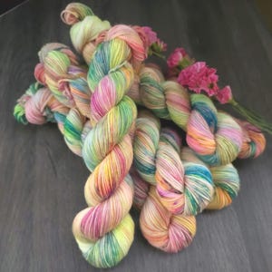 May include: A collection of colorful yarn skeins in shades of pink, yellow, green, and blue. The yarn is arranged with pink flowers on a dark wooden surface. The yarn appears to be hand-dyed, with a variegated color pattern.