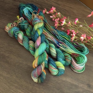 May include: A selection of hand-dyed yarn in vibrant hues of teal, green, yellow, and pink. The yarn is displayed with a sprig of pink flowers on a wooden surface. The yarn is suitable for knitting and crochet projects.