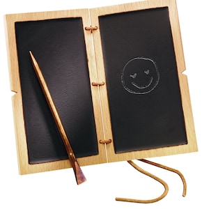 Può includere: Una lavagna in legno con una superficie di scrittura nera e uno stilo in ottone. La lavagna è chiusa e ha un cinturino in pelle. Un viso sorridente è disegnato sulla lavagna.