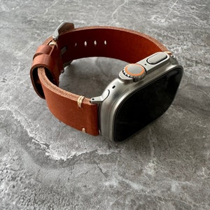 Pode incluir: Uma pulseira de relógio de couro marrom com um Apple Watch prateado com um mostrador preto. O relógio tem um botão prateado com um anel laranja ao redor.