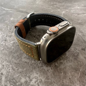 Puede incluir: Un Apple Watch plateado con una banda de lona verde y marrón. El reloj tiene una pantalla negra y una corona digital plateada con un anillo naranja.