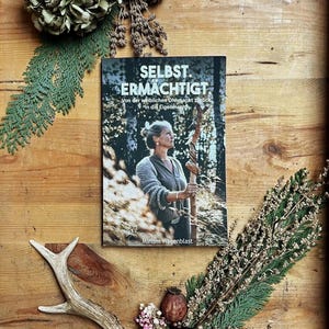 May include: A book titled "SELBST. ERMÄCHTIGT" with a woman holding a staff, surrounded by dried flowers and greenery on a wooden surface. The book cover features a woman in a forest setting. The author's name, Miriam Wasenblast, is visible.