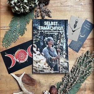 May include: A book titled "SELBST. ERMACHTIGT" with a woman on the cover, alongside a black card with "RUN WILD WOMAN" text and a crescent moon design. A necklace, a bookmark, and dried flowers are also present, all arranged on a wooden surface.