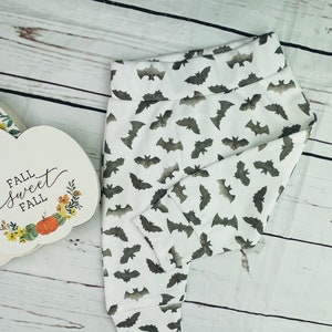 May include: White baby pants with a repeating pattern of gray bats. The pants have a folded waistband and fitted cuffs. A decorative pumpkin-shaped sign with the words "Fall Sweet Fall" is visible in the background.