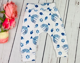 May include: White baby trousers featuring a blue pattern of menorahs, dreidels, and Stars of David. The trousers have a relaxed fit and are made of soft material. Ideal for Hanukkah celebrations.