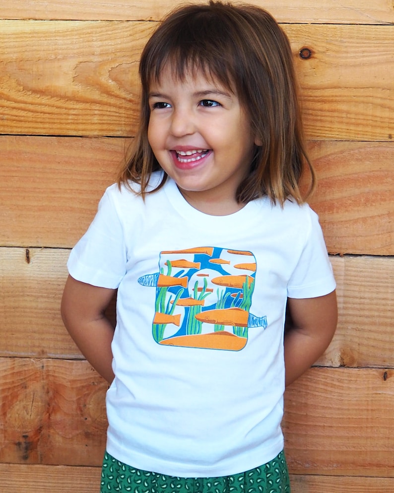 Puede incluir: Camiseta blanca con una ilustraci&oacute;n colorida de peces naranjas nadando en un entorno acu&aacute;tico azul y verde. La camiseta est&aacute; hecha de un material suave y c&oacute;modo, adecuada para ni&ntilde;os.