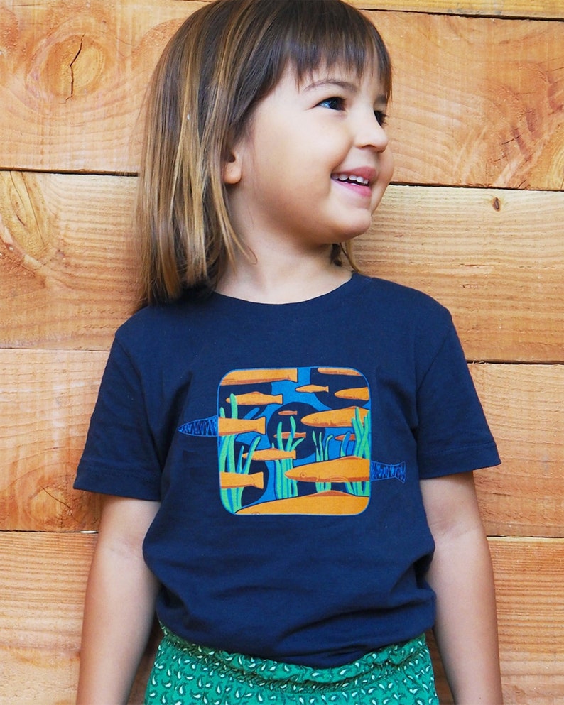 Puede incluir: Camiseta azul marino con un dise&ntilde;o cuadrado que muestra peces naranjas y algas verdes en una escena submarina azul. La camiseta tiene mangas cortas y la lleva un ni&ntilde;o.