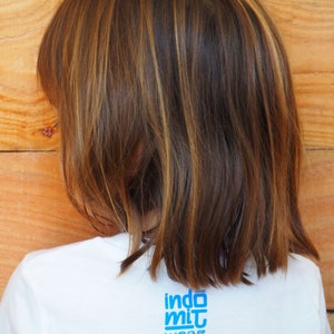Puede incluir: Vista trasera de una persona que lleva una camiseta blanca con el texto azul "indo mit wear". La persona tiene el pelo casta&ntilde;o con mechas rubias. El fondo es una pared de madera.