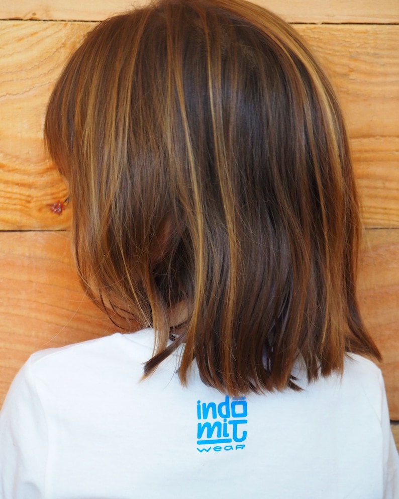 Puede incluir: Vista trasera de una persona que lleva una camiseta blanca con el texto azul "indo mit wear". La persona tiene el pelo casta&ntilde;o con mechas rubias. El fondo es una pared de madera.