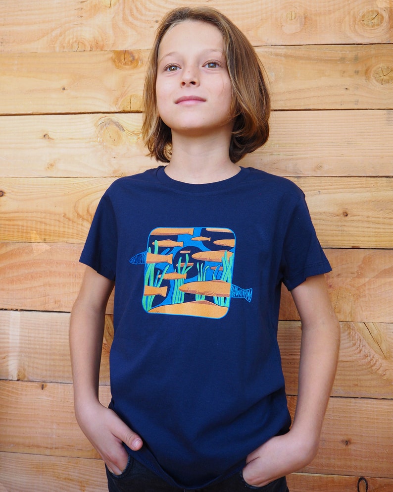 Puede incluir: Una camiseta azul marino con un gr&aacute;fico colorido de peces nadando en una escena submarina. El dise&ntilde;o incluye peces naranjas y azules, algas verdes y un fondo marino arenoso. La camiseta est&aacute; hecha de material suave.