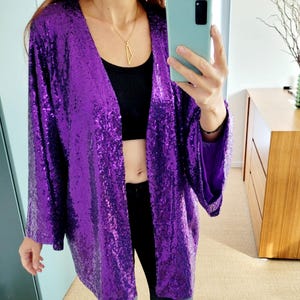 May include: A vibrant purple sequin jacket with long sleeves. The open-front jacket is paired with a black top and black pants. The outfit is completed with black boots. A gold necklace is visible.