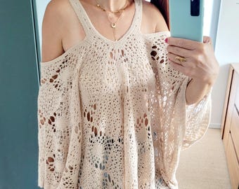 May include: A beige, open-shoulder crochet top with fringe detail. The top has a V-neck and a loose, flowing design. The sleeves are long and the shoulders are cut out. The top is paired with light blue jeans.