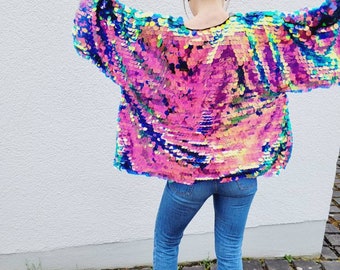 May include: A colorful sequin jacket with a black trim, paired with blue jeans and black boots. The jacket features a vibrant mix of pink, blue, and yellow sequins. The jeans have a frayed hem. The person is standing on a stone surface.