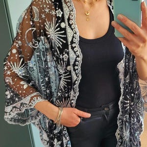 Peut inclure: Kimono noir transparent avec des sequins argentés et un motif brodé d'étoiles et de fleurs. Le kimono a un bord festonné et est porté sur un débardeur noir et un pantalon noir. Un collier et des bracelets dorés complètent le look.