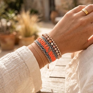 Könnte beinhalten: Ein Stapel aus drei Armbändern, bestehend aus einer dünnen Goldkette, einem gewebten Stoffarmband mit dem Text KARMA BABY in Orange und Schwarz sowie einem Perlenarmband mit hellrosa und goldfarbenen Kugeln, dazu ein goldener Ring am Finger.