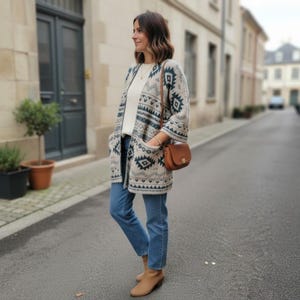 Könnte beinhalten: Eine beige, vorne offene Strickjacke mit dunkelblauem geometrischem Muster, getragen über einem weißen Oberteil und einer blauen Jeans. Das Outfit umfasst hellbraune Stiefeletten und eine kleine braune Umhängetasche aus Leder.