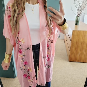 May include: A pink sheer kimono with floral embroidery. The kimono has short sleeves and a loose fit. The model is wearing the kimono over a white tank top and blue jeans.