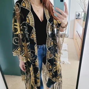 May include: A black sheer kimono featuring gold sequin embroidery in a fan and floral pattern. The garment has an open front, wide sleeves, and a long black fringe trim along the bottom hem. It is worn over a black top and blue denim jeans.