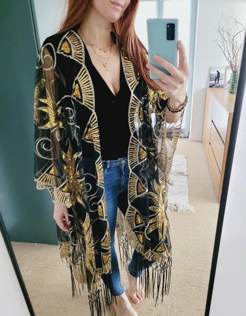 May include: A black sheer kimono featuring gold sequin embroidery in a fan and floral pattern. The garment has an open front, wide sleeves, and a long black fringe trim along the bottom hem. It is worn over a black top and blue denim jeans.