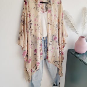 Könnte beinhalten: Ein durchsichtiges Kimono mit floralem Muster auf hellbeigem Hintergrund und rosa und violetten Blumen. Das Kimono ist über ein weißes Tanktop und eine blaue Jeans gelegt.