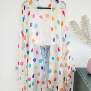 Könnte beinhalten: Weißer, durchsichtiger Kimono mit bunten Punkten und Quasten. Der Kimono ist über ein weißes Tanktop und eine blaue Jeans gelegt.