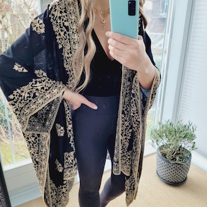 Könnte beinhalten: Ein schwarzer Kimono-Wrap mit aufwendiger goldener Stickerei. Der Wrap wird über einem schwarzen Oberteil und dunklen Leggings getragen. Das Bild wurde in Innenräumen aufgenommen, mit einer Topfpflanze im Hintergrund.