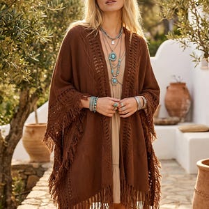 Puede incluir: Una mujer con un poncho marrón con flecos y diseño de ganchillo. Lleva múltiples collares plateados con colgantes turquesas y pulseras. El fondo incluye olivos y macetas de terracota, sugiriendo un estilo bohemio.