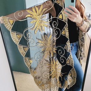 May include: A sheer black kimono featuring intricate gold sequin embroidery in floral and geometric patterns. The garment has wide, flowing sleeves, a long open front, and a bottom hem finished with delicate black fringe detailing.