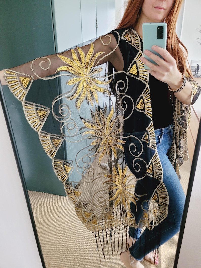 May include: A sheer black kimono featuring intricate gold sequin embroidery in floral and geometric patterns. The garment has wide, flowing sleeves, a long open front, and a bottom hem finished with delicate black fringe detailing.