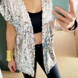 May include: Silver sequin vest with short sleeves. The open-front vest is paired with a black top and black pants. A gold necklace and gold bracelets complete the look. The outfit is suitable for parties and special events.