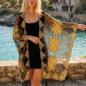 May include: A black sheer kimono featuring intricate gold embroidery in floral and geometric patterns. The garment has an open front, wide sleeves, and a long black fringe trim along the bottom hem, worn over a simple black sleeveless dress.