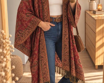 May include: A red and black paisley print open-front kimono featuring long wide sleeves and a fringed hemline. The garment is worn over a white ribbed tank top, blue denim jeans with a brown leather belt, and tan loafers, paired with a brown shoulder bag.