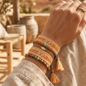 Könnte beinhalten: Ein Armbandstapel bestehend aus zwei gewebten Bändern mit dem Text GOOD KARMA in Orange, einer geflochtenen hellbraunen Lederschnur, einer dünnen Goldkette mit Hamsa-Anhänger und einer Reihe Holzperlen mit türkisfarbenen Akzenten und Quasten.