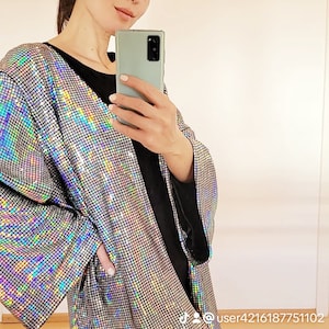 May include: A woman wearing a silver sequined jacket with iridescent rainbow hues. The jacket has a loose fit and long sleeves. The woman is wearing black leggings and holding a smartphone.