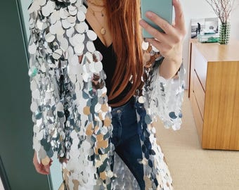 May include: A long, silver sequin hooded jacket. The jacket is made of many small, round, reflective sequins. The person in the image is wearing a black top and blue jeans. The jacket is open in the front.