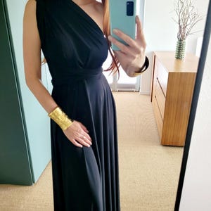 Peut inclure: Une robe longue noire à une épaule avec une jupe fluide. La robe est associée à un large bracelet manchette doré. L'image est prise devant un miroir.